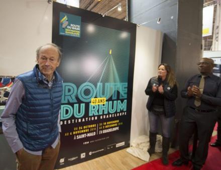 Mike Birch au dernier Nautic, aux côtés de l'affiche officielle de la prochaine édition. Ce dernier a gagné la première Route du Rhum en 1978 sur son trimaran jaune "Olympus Photo". Nautisme Article
