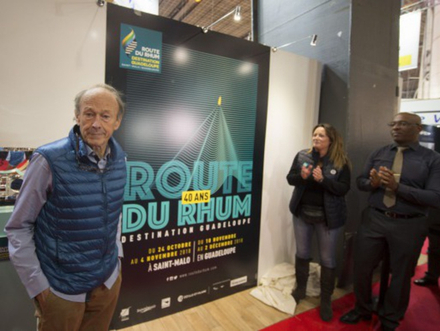 Mike Birch au dernier Nautic, aux côtés de l'affiche officielle de la prochaine édition. Ce dernier a gagné la première Route du Rhum en 1978 sur son trimaran jaune "Olympus Photo". Nautisme Article